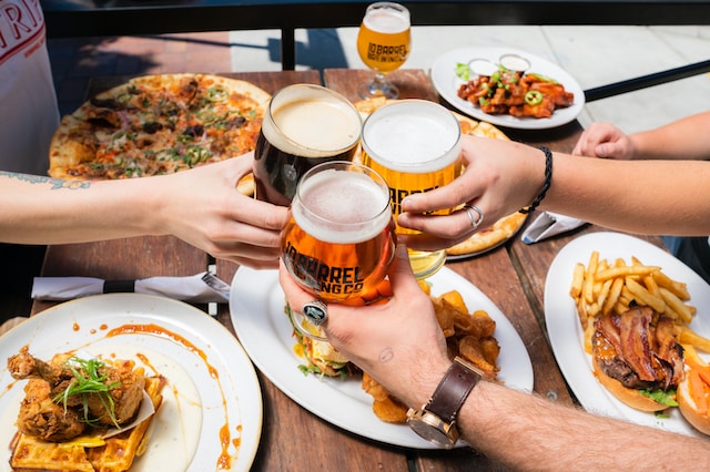 hands holding beer glasses in a toast over a table filled with food